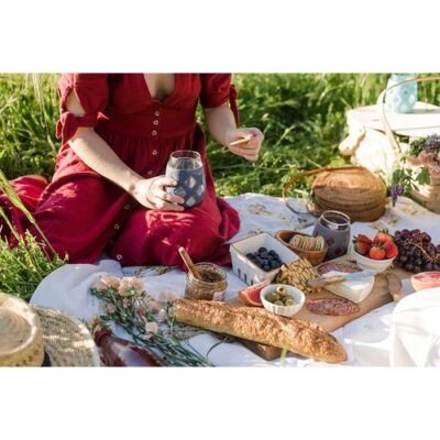 A woman in the picnic drinking wine in the wine tumbler and she kept variety of cheese items on the cheese board to enjoy her holiday