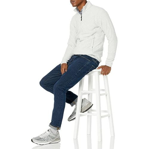 A young man wearing the quarter zip polar fleece jacket and blue plant with white shoes and sitting on the white stool